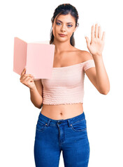 Poster - Young beautiful woman holding book with open hand doing stop sign with serious and confident expression, defense gesture