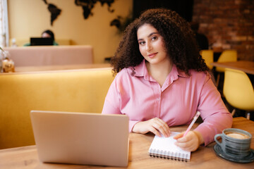 Beautiful stylishly dressed plump woman takes notes in a notebook. The interviewer is sitting at a laptop and a cup of coffee in a cafe during an interview. Meeting with a consultant in a cafe.