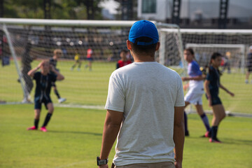 Father standing and watching his daughter playing football in a school tournament on a clear sky and sunny day. Sport, outdoor active, lifestyle, happy family and soccer mom and soccer dad concept.