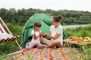 Wall Mural - Photo of woman with bun hairstyle wearing white t shirt sitting near the tent near the river with her daughter and drinking coffee or tea, family having camping, resting.