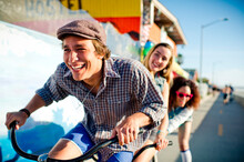 Young Man Smiling As He Pedals Tandem With Two Young Women Behind Him On The Boardwalk At The Beach