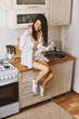 © olga pink - Asian brunette woman in white in kitchen. Female holding, drinking morning cup hot coffee tea