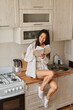 © olga pink - Asian brunette woman in white in kitchen. Female holding, drinking morning cup hot coffee tea