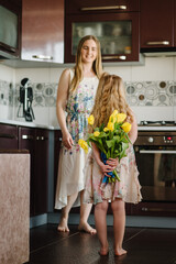 Wall Mural - Happy mother's day. Child daughter congratulates the mom and gives her bouquet of flowers tulips in kitchen. Mom and girl smiling. Family holiday and togetherness. Back view. International Women's Day