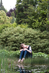 Wall Mural - melancholic young woman with long blond hair seating on a bank of a pond in summer park