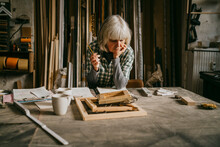 Thoughtful Senior Female Carpenter Holding Pencil While Leaning On Workbench