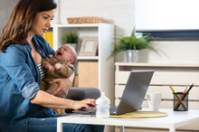 Portrait Of Casual Young Business Mom Holding Her Cute Newborn Baby While Working On A Laptop Computer At The Home Office.  Working Mother Using Computer And Nursing Newborn Child