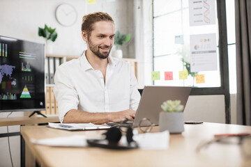 Wall Mural - Busy caucasian man in white shirt sitting at office and using wireless laptop. Big monitor with various charts and graphs on background. Business and finance concept.