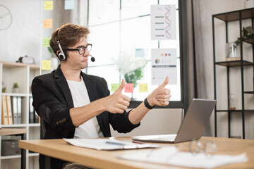 Young businessman in headset using laptop computer during video call working in office. Concentrated adult successful man wearing official suit sitting at wooden desk indoor showing thumbs up