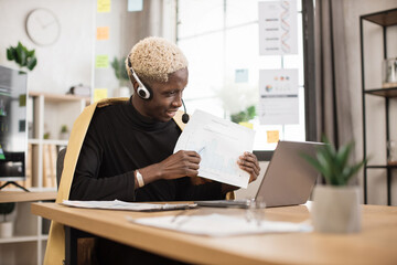 Wall Mural - Serious marketing african man in suit and headset talking to client on video call, presenting paper graphic reports, showing, explaining diagram. Employee, manager attending online meeting