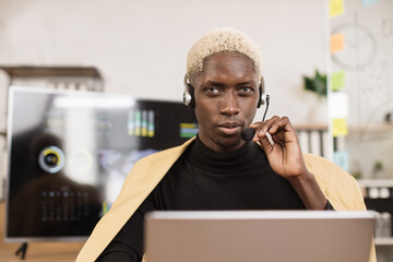 Wall Mural - Confident african man in yellow suit using headset sitting at modern office and using wireless laptop. Huge monitor with various charts and graphs on background. Business finance technology concept