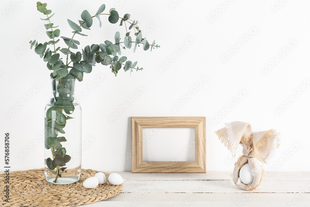 Blank frame, vase with eucalyptus and Easter eggs on table near white wall
