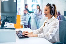 Colorful Photo Of Female IT Sector Operator With Headset Talking On Microphone During Online Support Call And Using Computer At Office.