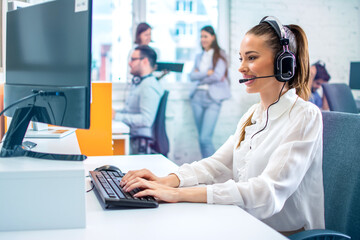 Colorful photo of female IT sector operator with headset talking on microphone during online support call and using computer at office.