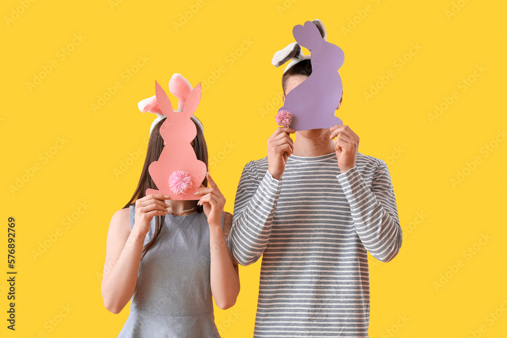 Happy young couple in bunny ears with paper Easter rabbits on yellow background