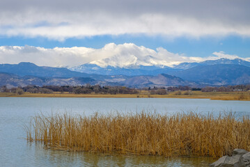 View of lake; mountain range and peak with snow behind it; cloudy sky in background with clouds descending on the peak in winter