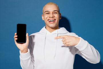 Wall Mural - Young dyed blond man of African American ethnicity in white hoody hold use point finger on mobile cell phone with blank screen workspace area isolated on plain dark royal navy blue background studio