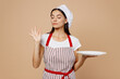 Young housewife housekeeper chef baker latin woman wear striped apron toque hat hold empty plate making okay taste delight sign savoring delicious isolated on plain beige background Cook food concept