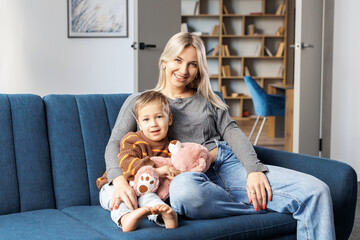 Young pregnant woman and her son sit on sofa at home, little boy spend time together, enjoy communication feeling kicks of unborn baby.