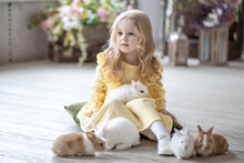 A Little Smiling Girl In A Yellow Dress Is Playing On The Floor In A Room With Rabbits. A Baby And A Rabbit. The Living Room Is Decorated For Easter.