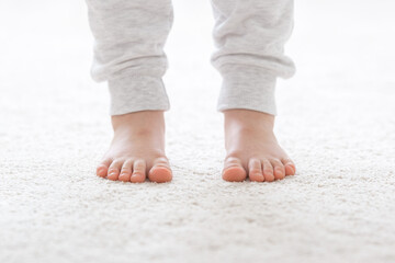 Little child legs in white warm trousers standing on light beige carpet at home room. Barefoot closeup. Front view.