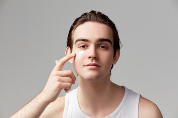 Portrait of young handsome boy applying moisturizing face cream, mask, posing against grey studio background. Concept of men's health, body and skin care, hygiene and male cosmetology