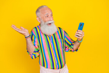 Photo of positive optimistic retired man with white beard dressed striped shirt hold smartphone isolated on yellow color background