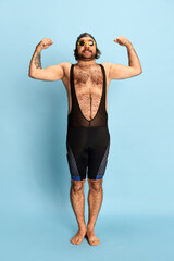 Comic portrait of funny bearded man in swimsuit and swimming cap and goggles having fun isolated over blue background. Concept of sport, active lifestyle and humor