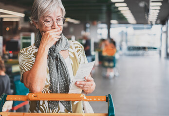 Wall Mural - Senior woman in the supermarket checks her grocery receipt looking worried about rising costs - elderly lady pushing shopping cart, consumerism concept, rising prices, inflation