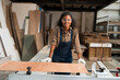 Portrait of young female carpenter in wood workshop. Happy, Smiling black female joiner wearing safety glasses in workshop at wooden warehouse. SME, Start up and small business concept