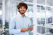 © Liubomir - Portrait of young programmer with tablet computer inside office, hispanic man with curly hair smiling and looking at camera, man at workplace testing online applications standing near window.