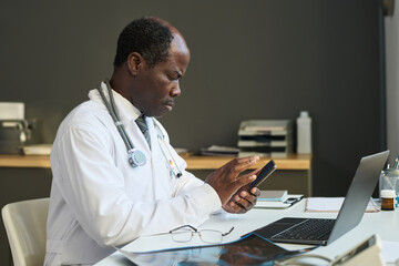 Poster - Side view of serious mature clinician scrolling in smartphone while sitting in front of laptop by workplace and consulting online patients