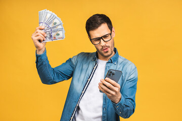 Happy winner! Excited man in casual holding lots of money in dollar currencys and using phone in hands isolated over yellow background.