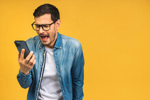 Young Caucasian Man Angry, Frustrated And Furious With His Mobile Phone, Angry With Customer Service. Isolated Over Yellow Background.