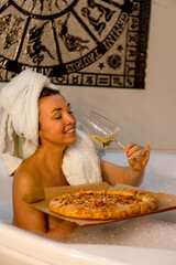 portrait of an attractive dark-haired girl with a towel on her head .the girl sits in the bath drinks wine and eats pizza