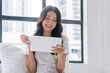 © kaew6566 - Positivity asian woman holding digital tablet sitting with pillow white bed beside window at home. Happy young female sitting relax looking something on tablet social media at home. Technology concept