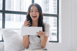 © kaew6566 - Positivity asian woman holding digital tablet sitting with pillow white bed beside window at home. Happy young female sitting relax looking something on tablet social media at home. Technology concept