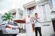 Asian Muslim couple with hand gestures saying Eid al-Fitr standing in front of a house with a car