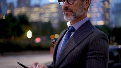 Mature successful man in suit and stylish eye glasses typing text message on smartphone device while standing near city road with with passing car traffic. Businessman browsing network on cellular app