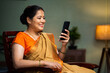© WESTOCK - Middle woman in sare buy using mobile phone on chair at home - concept of technology, social media sharing, internet.