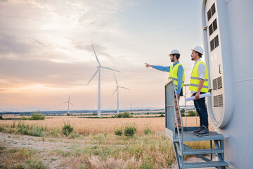 Two wind mill engineers or technicians planning a construction indicating where the new turbine will be placed. A couple of renewable energy workers wearing hardhat or helmet and reflective vests