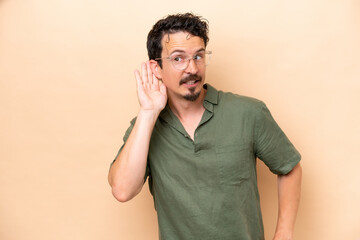 Wall Mural - Young caucasian man isolated on beige background listening to something by putting hand on the ear