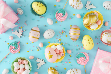 Wall Mural - Easter celebration concept. Top view photo of color eggs, easter candy and chocolate sprinkles with meringue cake toppers and gift boxes on pastel blue background. Easter decoration idea.