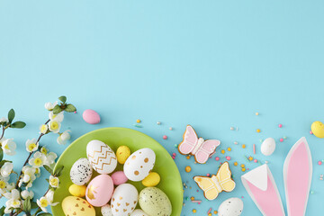Wall Mural - Easter decoration concept. Top view composition of green plate with colorful eggs easter bunny ears gingerbread sprinkles and cherry blossom branch on isolated pastel blue background with empty space