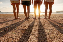 Group Of Friends Stands At Sunset Sea Beach. Closeup Photo Of Legs And Long Shadows