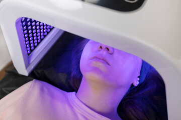 Blue led light mask for face skin therapy, care. Young girl gets problem skin treatment, lying on couch under blue light mask. Stimulation of collagen production, killing acne bacteria. Horizontal.