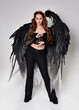 Full length portrait of beautiful woman with long red hair wearing sheer corset top, leather pants, large black angel feather wings. Standing pose, walking forwards with gestural hands reaching out. I