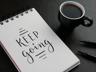 keep going letternig in notebook with cup of coffee and pens on black wooden desk