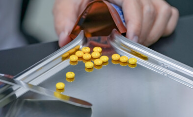 Selective focus on yellow tablets pills on stainless tray with blur hand of pharmacist or pharmacy technician counting pills into a plastic zipper bag. Prescription medicine. Medical health care.