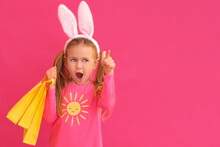 Easter Shopping. Excited Emotion Surprise Adorable Child Girl In Bunny Ears Rabbit Costume Hold Yellow Shopping Bags Isolated Pink Background With Finger Pointed Up. Kid Sale Discounts Easter Children